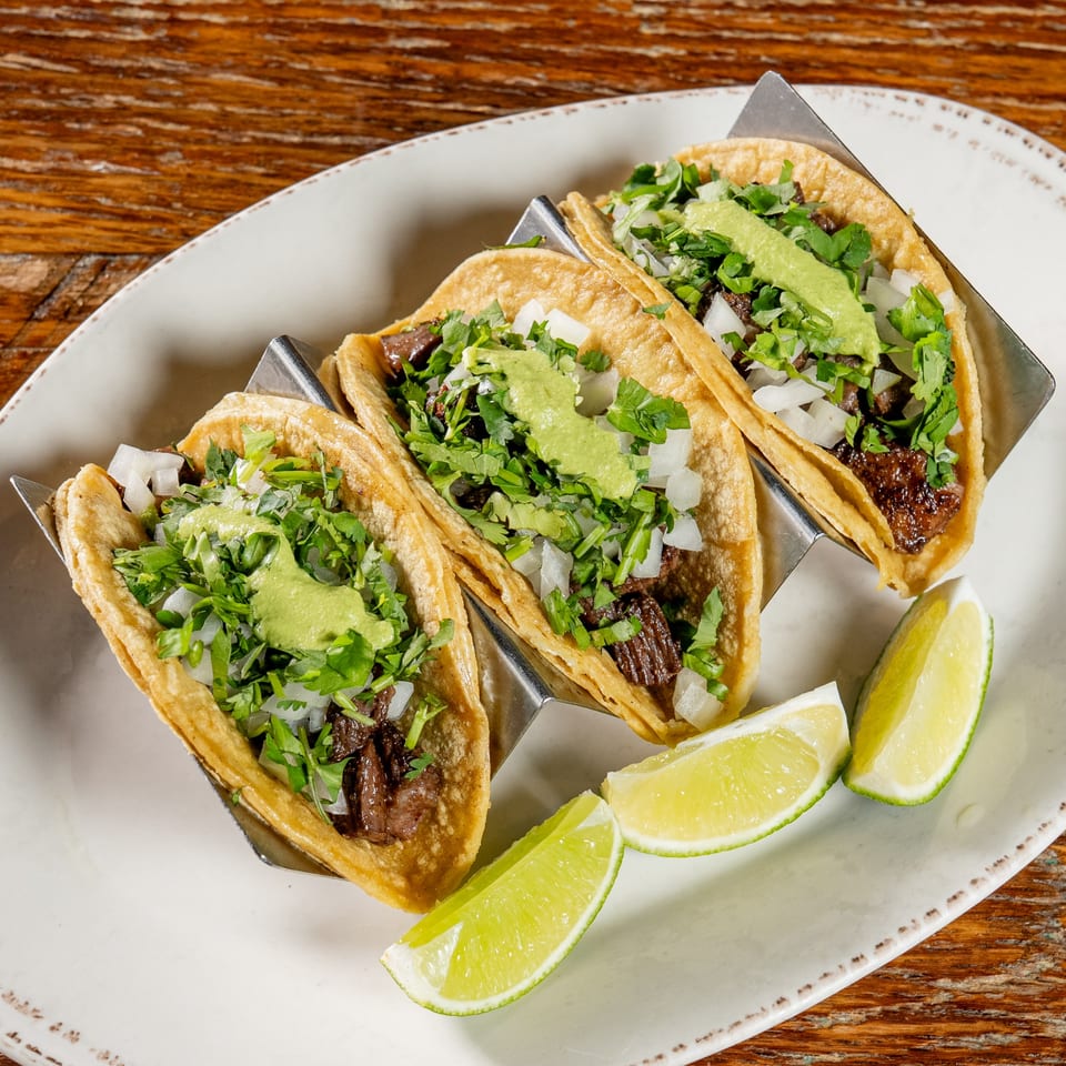 Three El Camino Mexican tacos with meat, cilantro, onions, green sauce, and lime wedges on a white plate.