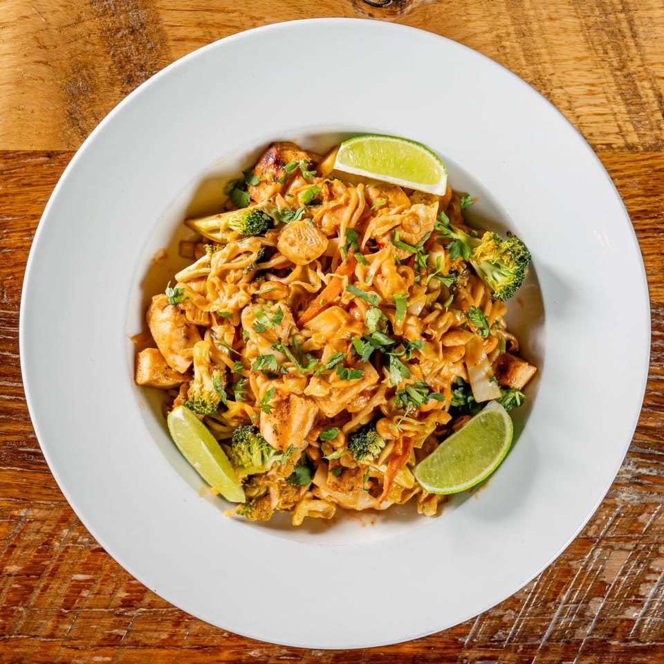 A white bowl of chicken and broccoli pasta with lime wedges on a wooden table.