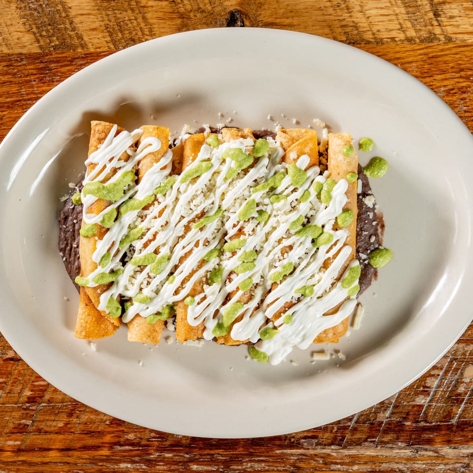 Crispy taquitos drizzled with white sour cream, green sauce, and crumbled cheese on a bed of refried beans.