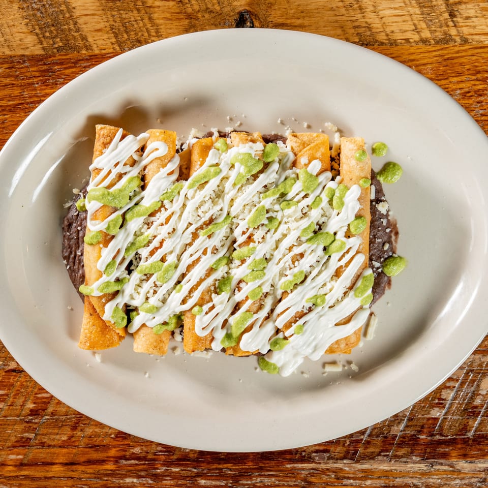 Crispy El Camino taquitos drizzled with white crema, green sauce, and cotija cheese on a bed of refried beans.